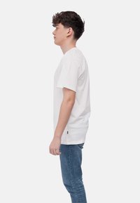 T-shirt bianco in cotone con scollo rotondo classico, maniche corte e vestibilità rilassata, abbinato a jeans blu. Accento del marchio discreto sull'orlo.
