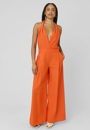 Femme portant une combinaison orange à col halter et jambes larges avec un décolleté profond, debout, une main dans la poche et les cheveux tressés.