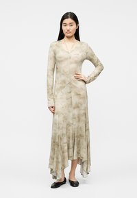 Nő áll hosszú ujjú bézs virágos ruhában, amelynek aszimmetrikus az alja, fekete lapos cipővel, egyik kezét a csípőjén tartva.