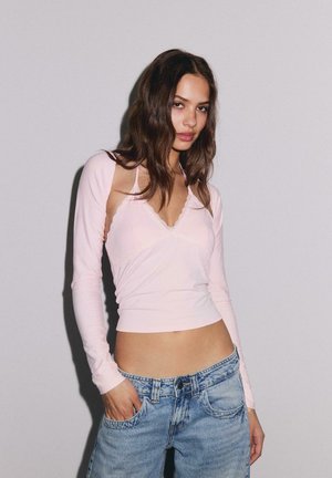 Junge Frau mit langen dunklen Haaren, die ein blassrosa, langärmeliges, bauchfreies Oberteil und locker sitzende blaue Jeans trägt, steht vor einem schlichten hellen Hintergrund.