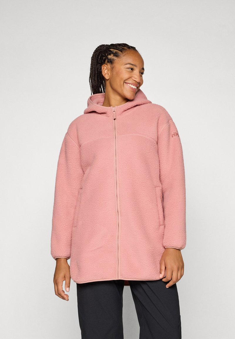 Donna sorridente con una giacca in pile rosa con cappuccio, in piedi su uno sfondo grigio chiaro. Ha i capelli intrecciati e indossa pantaloni casual.