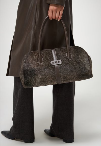 Borsa a mano in feltro marrone con dettagli in pelle, forma ovale, doppio manico e dettaglio del logo in metallo. Indossata con un cappotto marrone e pantaloni.