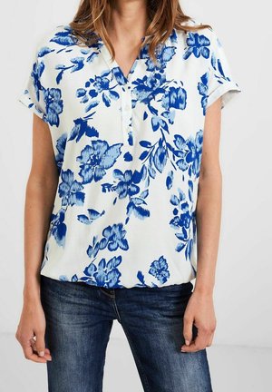Femme portant un chemisier blanc à manches courtes avec un imprimé floral bleu et un jean bleu foncé, debout devant un fond clair uni.