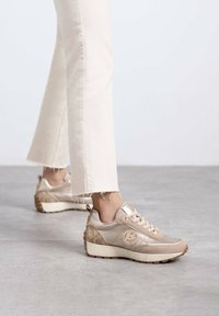 Zapatillas beige con un acabado metálico, acentos texturizados y un logo. Combinadas con unos jeans blancos cortos con dobladillos deshilachados.