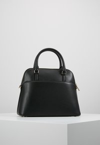 Černá kožená kabelka s kulatými uchy a zlatým zipem, vystavená na bílém podstavci na jednoduchém šedém pozadí.