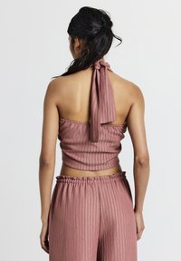 Donna con i capelli scuri raccolti, che indossa un top halter a coste color rosa polveroso e pantaloni a vita alta abbinati, di spalle su uno sfondo neutro.