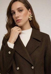 Ruskea kaksirivinen takki, jossa suuret hopeanväriset napit, päällä valkoinen ribbineulottu pooloneule. Malli esittelee näyttävät kultaiset korvakorut.