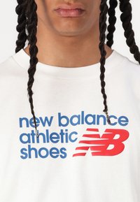 Fehér pamut póló, amelyen kék szöveg olvasható: "new balance athletic shoes", valamint egy piros New Balance logó. Kerek nyakkivágású és rövid ujjú.