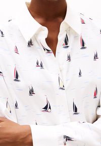 Chemise blanche à boutons avec un motif de voilier en noir, rouge et jaune, avec un col classique et une texture de tissu lisse.
