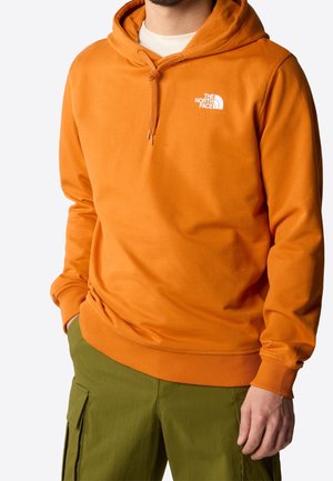 Mężczyzna w jaskrawo pomarańczowej bluzie z kapturem The North Face i oliwkowych spodniach typu cargo, z jedną ręką w kieszeni, kadrowany od szyi do połowy uda.