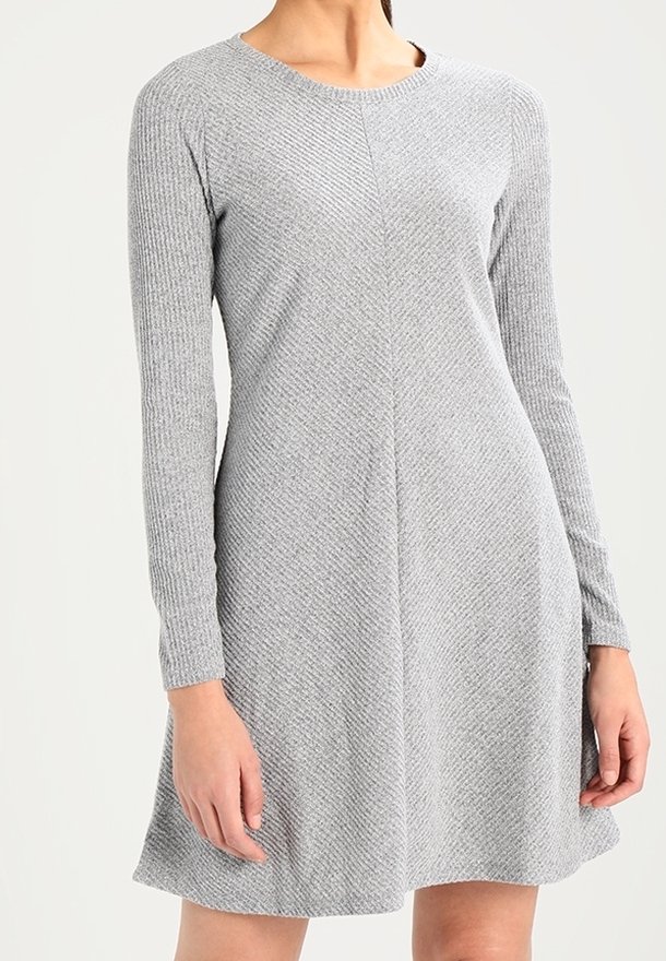 Femme portant une robe en maille texturée gris clair à manches longues, avec un col rond et une jupe évasée, debout devant un fond uni.