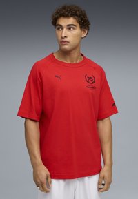 Rød bomulds T-shirt med korte ærmer, med et marineblåt logo og tekstprint på fronten. Enkel, løs pasform.