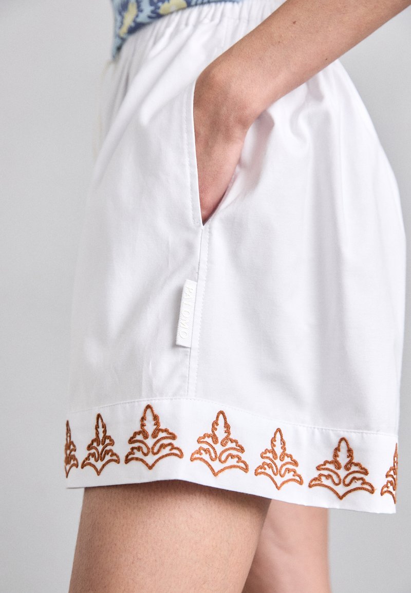 White shorts with side pockets and a decorative orange embroidered pattern along the hem. Tag with "PALOMO" visible on the waistband.