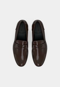 Καφέ δερμάτινα μοκασίνια με κομψό σχεδιασμό, ομαλή υφή, ραφές κατά μήκος της άκρης και μια διακριτική ανυψωμένη ταινία στην κορυφή.