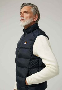 Chaleco sin mangas acolchado de color azul marino con acabado mate, cuello alto y bolsillos laterales. Lleva puesto un suéter de punto blanco texturizado.