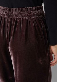 Paarse fluwelen broek met een gerimpelde elastische tailleband, met een zachte textuur en een losse pasvorm, zijaanzicht weergegeven.