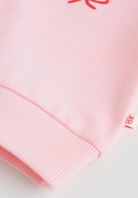 Różowa rękawica bluzy z ribbowanym mankietem i małą beżową metką z ikoną czerwonego pudełka na prezent na białym tle.