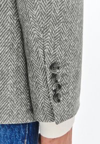 Tkanina żakietowa w szary wzór w jodełkę z czterema czarnymi guzikami na mankiecie i kremowymi szczegółami dzianiny przy otwarciu rękawa.