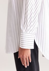 Hand hangt aan de zijde, gekleed in een wit shirt met dunne verticale zwarte strepen en donkere broek, getoond van elleboog tot bovenbeen.