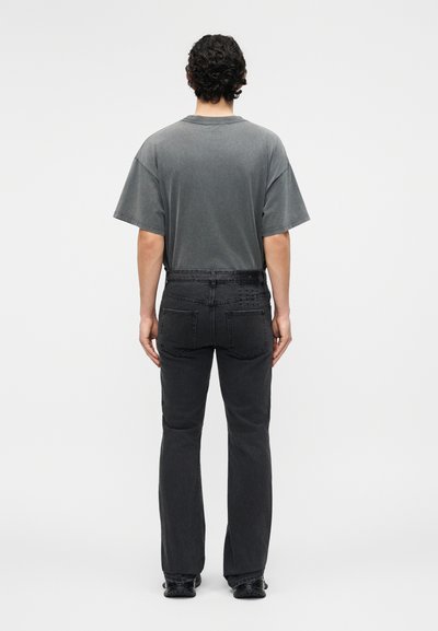 Άνδρας με σγουρά σκούρα μαλλιά στέκεται με την πλάτη γυρισμένη, φοράει μια ξεθωριασμένη γκρι υπερμεγέθη μπλούζα και μαύρο τζιν ίσιας γραμμής με μαύρα παπούτσια.