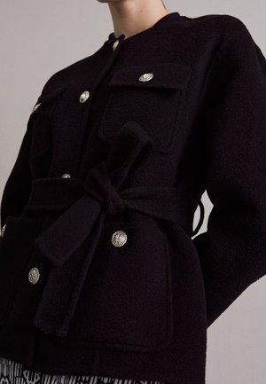 Nærbillede af en sort tekstureret jakke med sølvfarvede dekorative knapper, to brystlommer, et bundet bælte i taljen og lange ærmer.