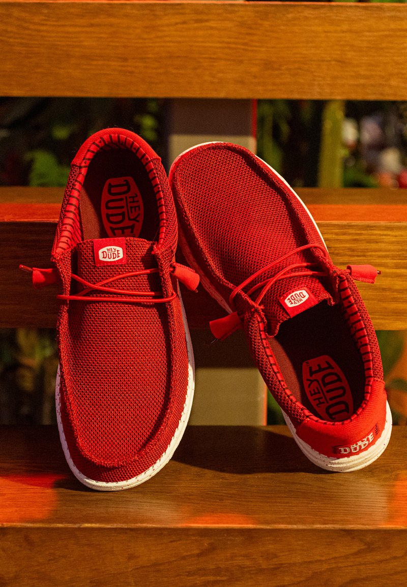 Coppia di sneakers casual rosse slip-on con suole bianche posate su una panchina di legno, con marchio "Hey Dude" all’interno e sulla linguetta.