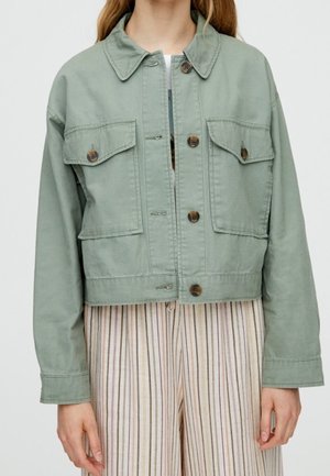 Person trägt eine hellgrüne, mit Knöpfen versehenen Jacke mit zwei Brusttaschen und beige gestreifte Hose.