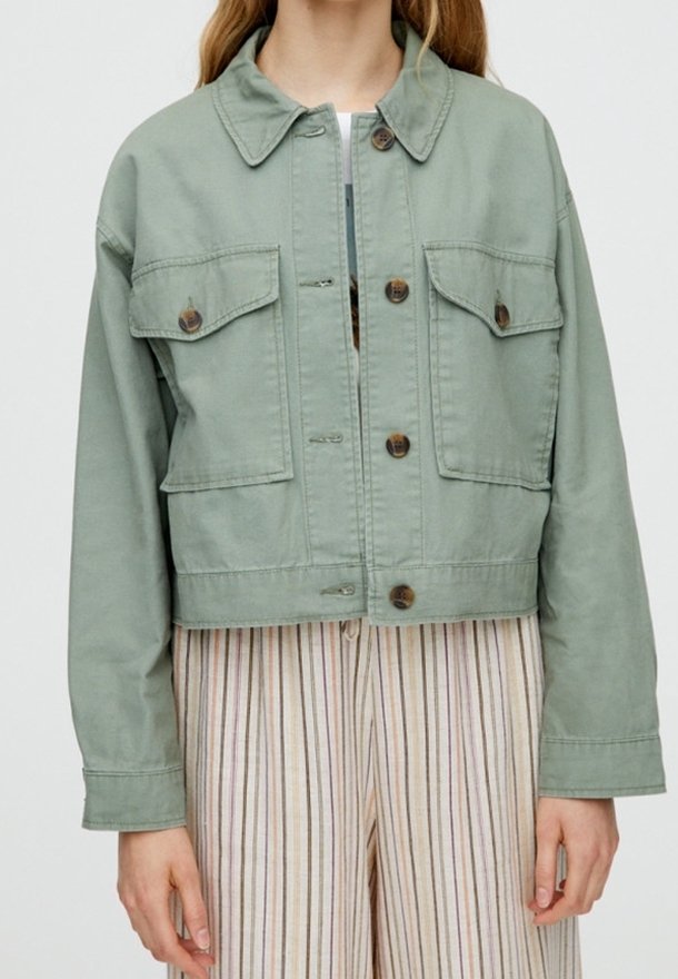 Personne portant une veste boutonnée vert clair avec deux poches poitrine avant et un pantalon beige à rayures.