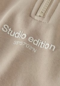 Tejido beige con texto en relieve blanco: "Edición de estudio" con coordenadas "55°57'03"N" debajo; una cremallera parcial en la parte superior.