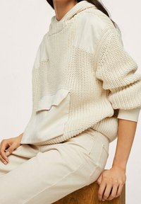 Kremowa dzianinowa bluza z kapturem z fakturowanym splotem, kontrastującymi gładkimi panelami i przednią kieszenią; noszona z beżowymi spodniami w tym samym odcieniu.