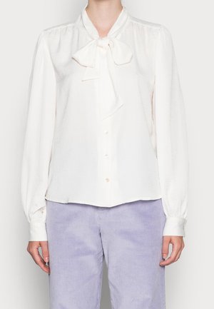 White long-sleeve blouse with front buttons and tied bow collar paired with light purple corduroy pants worn by a standing person.