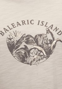 Hellbeige Textil mit einem kreisförmigen Design, das dunkle Blattillustrationen und den Text "BALEARISCHE INSEL" in kräftigen, kontrastierenden Farben zeigt.