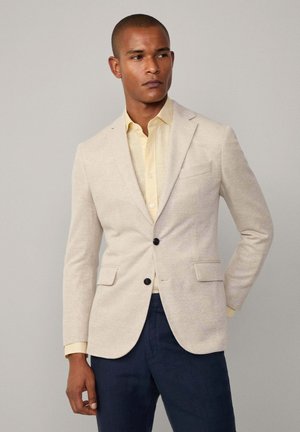 Beige Leinenmischung-Blazer mit Reverskragen, zwei Fronttaschen und dunklen Knöpfen, über einem hellgelben gestreiften Hemd getragen.