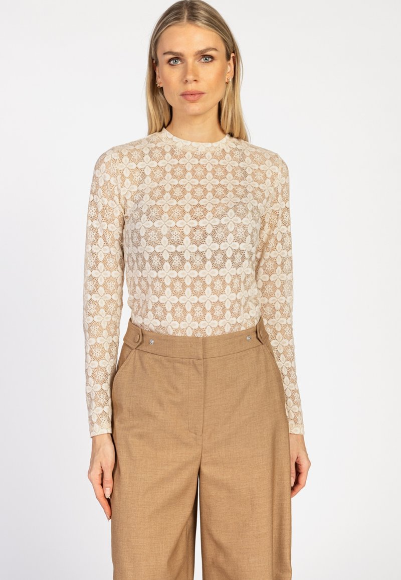 Blonde vrouw in een beige bloemenkant lange mouwen top, gestoken in hoge taille camel kleurige wijde broek op een witte achtergrond.