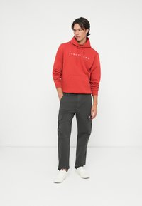 Červená mikina s kapucňou a predným vreckom, ozdobená bielym logom; v kombinácii so šedými cargo nohavicami s bočnými vreckami; doplnené bielymi teniskami.