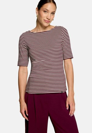 Femme portant un haut à manches courtes rayé bordeaux et blanc et un pantalon bordeaux, debout devant un fond clair uni.