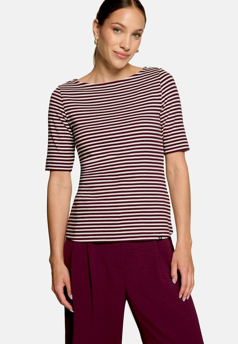 Femme portant un haut à manches courtes rayé bordeaux et blanc et un pantalon bordeaux, debout devant un fond clair uni.