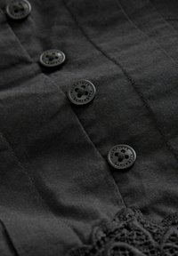 Camisa de tela negra con tapeta de botones y botones redondos texturizados, además de un intrincado detalle de encaje en el dobladillo.