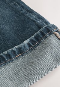 Šev dámskych riflí ukazujúci tmavú pranú časť navrchu a svetlú pod ňou, so strapcovaným okrajom a kontrastným prešitím. Mäkká, textúrovaná látka.