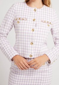 Chaqueta a cuadros lavanda en tejido texturizado, con botones dorados y detalles de cadena, con un escote redondo y dos bolsillos en el pecho.