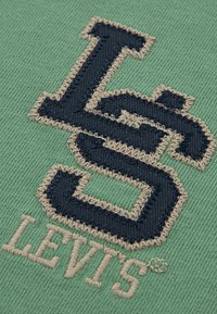 Zelená látka s textúrovaným povrchom, na ktorom sa nachádza výrazné vyšívané logo "L" a "5" v námorníckej modrej farbe, doplnené béžovým prešívaním.