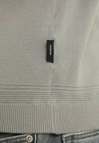 Pull gris en tricot avec ourlet et poignets côtelés. Présente une étiquette de marque noire cousue sur le côté. La texture est lisse et légère.