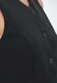 Κοντινό πλάνο μαύρου αμάνικου ενδύματος με τρία ορατά κουμπιά στο εμπρός μέρος και λεία υφή υφάσματος.