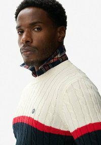 Maglione a treccia lavorato a maglia in crema, rosso e blu navy. Presenta un colletto a camicia a quadri sovrapposto. Texture morbida e maglia a coste.