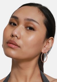 Boucles d'oreilles créoles en or rose avec une texture frappée, de grande forme ronde et une fermeture sécurisée, présentées sur l'oreille d'un modèle.