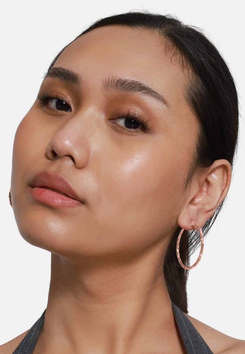 Boucles d'oreilles créoles en or rose avec une texture frappée, de grande forme ronde et une fermeture sécurisée, présentées sur l'oreille d'un modèle.