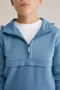 Světle modrá fleecová mikina s kapucí a designem na polovinu zipu, texturovaným spodním panelem a vyšívaným logem. Obsahuje kapuci a hladký límec.