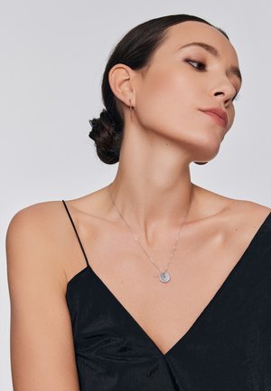 Srebrny naszyjnik z okrągłym zawieszką i akcentem w formie pasku, noszony na czarnej, półprzezroczystej bluzce z cienkimi ramiączkami, podkreślającej gładkie tekstury.