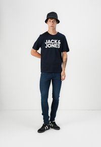 Jūrų kariuomenės medvilnės marškinėliai su balta "JACK & JONES" logotipu, derinami su mėlynomis džinsais ir mustais sportiniais bateliais, uždedami juodu kibiro skrybėle.