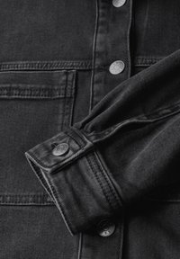 Musta farkkupaita, jossa on metalliset napit, käännettävä kaulus ja teksturoidut kankaat, joissa on näkyvä ompelu ja taskuyksityiskohta.